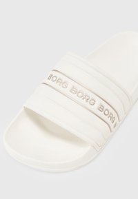 Sandale blanche à glissière avec trois bandes horizontales embossées portant la mention "BORG." Texture douce avec une semelle intérieure lisse et courbée. Design minimaliste.