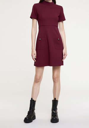 Femme portant une robe bordeaux à manches courtes avec des poches avant et des bottes de combat noires à lacets, debout devant un fond uni.