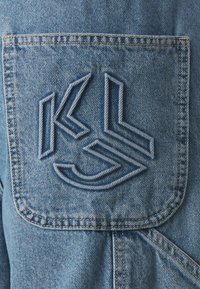 Tissu en denim de couleur bleu clair avec un logo brodé en bleu foncé sur une poche arrière. Surface texturée avec des détails de couture visibles.