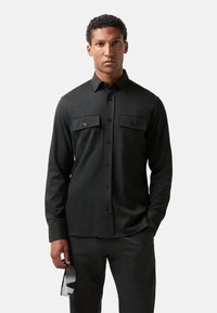 Dark grey long-sleeve shirt with two chest pockets, button-up front, and textured fabric. Styled with matching pants and sunglasses.