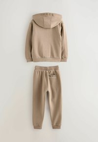 Beige kapuutsiga pusakas ja dressipükside komplekt, mis on valmistatud pehmest kangast. Kapuusil on seljal kõrge "BAKER" logo; pükstel on ka logo vööl.