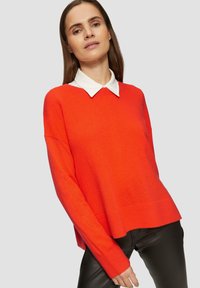 Roter, locker sitzender Pullover mit einem weißen Hemd darunter. Stricktextur, gerippte Bündchen und Saum sowie überschnittene Schultern.