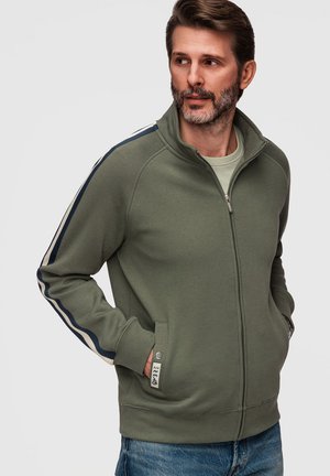 Man met baard die een olijfgroene jas met ritssluiting draagt met blauwe en beige strepen op de mouwen, staand met handen in de zakken, kijkt naar rechts.