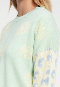 Ανοιχτοπράσινο πουλόβερ με απαλές κίτρινες μαργαρίτες στο σώμα και κρεμ μανίκια με ακανόνιστα γαλάζια στίγματα, φορεμένο από ένα άτομο.