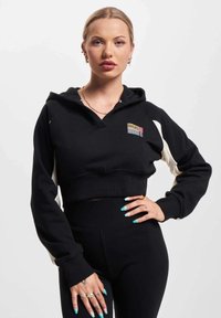 Črn skrajšan hooded pulover z belimi rokavi, ki vsebuje značko z logotipom. Material se zdi mehak in teksturiran. Model nosi črne legice.