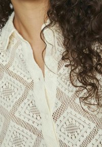 Blouse blanche en dentelle avec un motif en losange, dotée d'une fermeture à boutons et d'un col. Le tissu présente une apparence délicate et texturée.