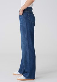 Jean en denim à jambes larges de couleur bleu foncé, avec une taille haute, deux poches latérales et une texture lisse. Assorti avec des baskets blanches.