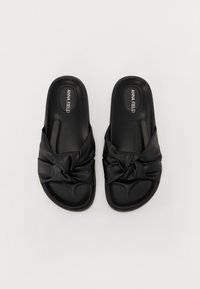 Black slide sandals made from smooth material, featuring a knotted design across the upper. Flat sole with textured detailing.