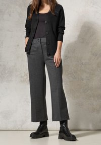 Schwarze gerippte Strickjacke über einem schwarzen Top, kombiniert mit weiten grauen Hosen und schwarzen Knöchelstiefeln mit robusten Sohlen. Geschmeidige Texturen und maßgeschneiderte Passform.