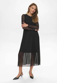 Rochie neagră cu mâneci lungi, realizată dintr-un material transparent cu model buline, design plisat și o siluetă fluidă. Asortată cu tocuri subțiri negre.