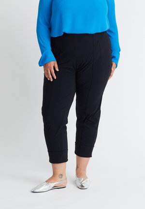 Personne portant un pantalon noir raccourci, un haut bleu à manches longues, des chaussures plates argentées, avec des tatouages à la cheville, debout devant un fond uni.