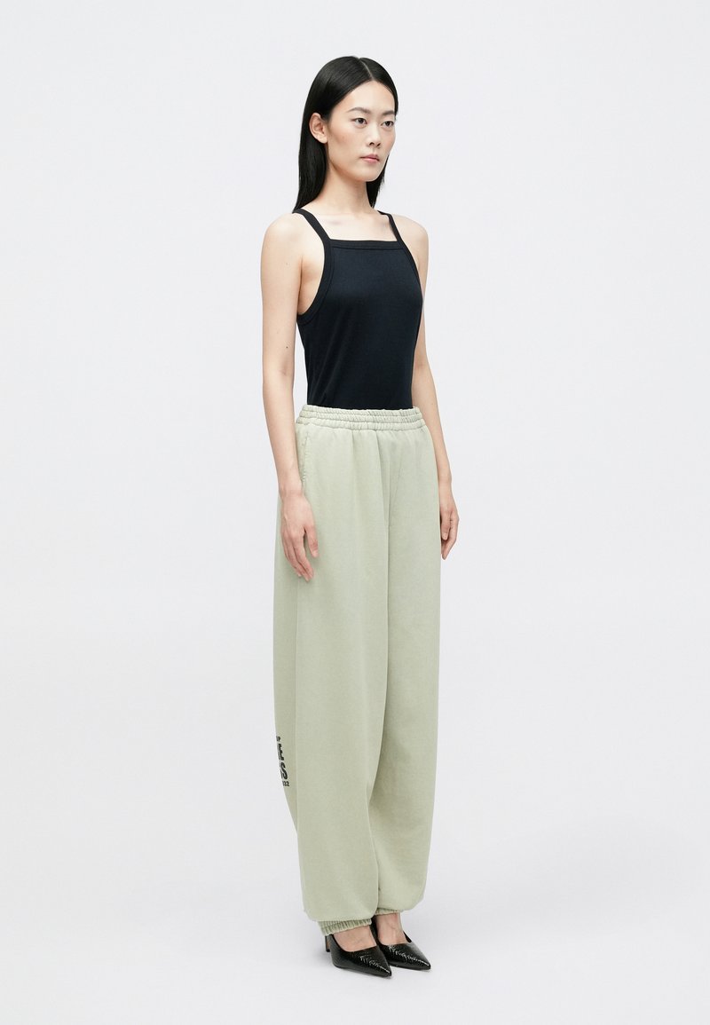 Zwarte strakke tanktop, lichtgroene, ruime sweatpants met elastiche tailleband en logo-detail, gecombineerd met zwarte puntige hakken.