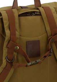 Sac à dos en toile beige avec des accents en cuir marron, quincaillerie en métal, bretelles ajustables et un patch cousu représentant un design d'ancre.