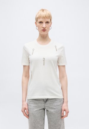 Rövid szőke hajú nő fehér pólót visel, amelyen öt ezüst és láncos díszítés található, világosszürke farmerrel, egyszínű háttér előtt áll.