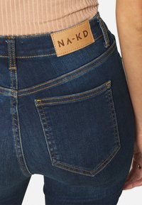 Unos pantalones vaqueros de mezclilla azul oscuro presentan una cintura alta, costuras naranjas y un parche de cuero con el logo "NA-KD" en la parte trasera. Se pueden ver dos bolsillos traseros.
