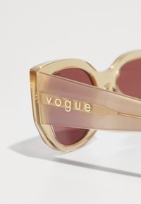 Ochelari de soare cu un cadru transparent de culoare maro deschis și lentile roșii. Au un logo auriu pe lateral. Formă unghiulară distinctivă și finisaj lucios.