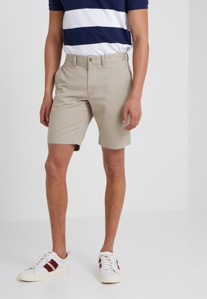 Homme portant un short beige au genou, un t-shirt rayé bleu marine et blanc et des baskets blanches avec des rayures bordeaux, debout sur un sol blanc.