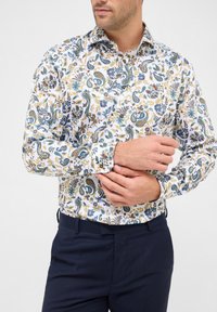 Man die de manchetten van een wit shirt met een blauw, groen en geel bloemenpaisleypatroon aanpast, in combinatie met donkerblauwe broek.