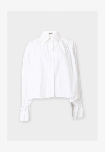 Blouse blanche à manches longues avec poignets froncés, boutons sur le devant, col pointu et coupe ample, présentée sur un fond uni.