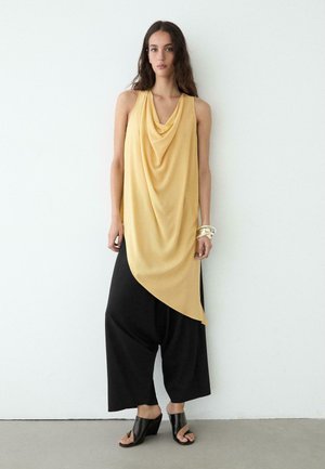 Junge Frau trägt ein ärmelloses, gelbes, drapiertes Oberteil, weite schwarze Hosen, schwarze offene Schuhe und silberne Armbänder, steht vor einer weißen Wand.