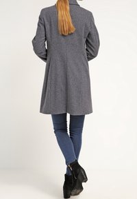 Grå ullkappa med skräddarsydd passform, notch-krage och ryggslits, kombinerad med blå jeans och svarta ankelboots. Slät textur, knälång.