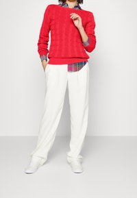 Röd kabelstickad tröja över en blå rutig skjorta, kombinerad med vita vida byxor och vita sneakers. Mjuka texturer, avslappnad stil.