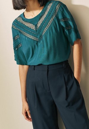 Teal blouse with sheer lace panels on chest and sleeves paired with high-waisted black trousers, hand resting in pocket against beige wall.