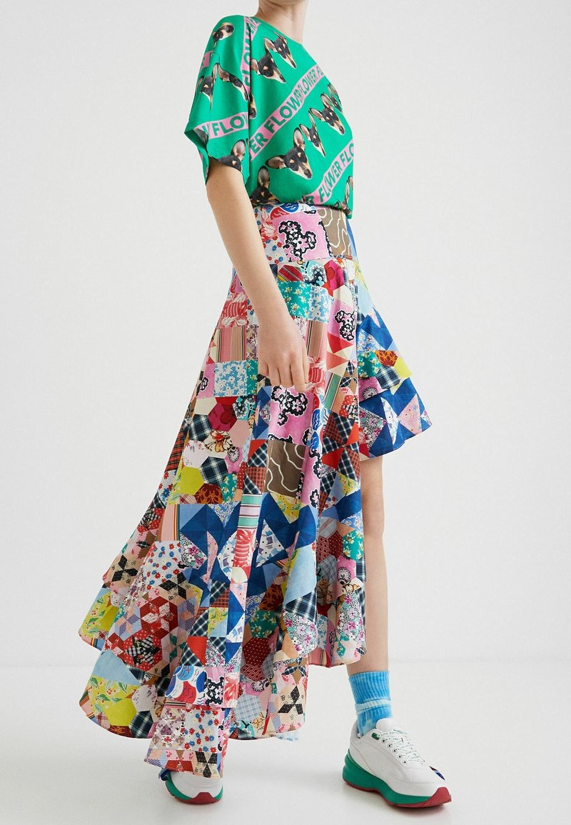Personne portant une chemise verte à motif chien associée à une jupe asymétrique colorée en patchwork et des baskets blanches avec des chaussettes bleues.