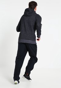 Uomo che si allontana indossando una felpa con cappuccio grigio scuro, pantaloni sportivi neri e scarpe da corsa nere su uno sfondo bianco.