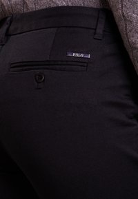 Schwarze maßgeschneiderte Hose mit strukturiertem Finish, ausgestattet mit einer geknöpften Gesäßtasche und einem metallischen Logo-Element am Bund.