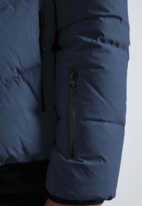Mørkeblå jakke med dun, som har struktur i stoffet, med en sort lynlåslomme og et quiltet mønster for isolering og stil.