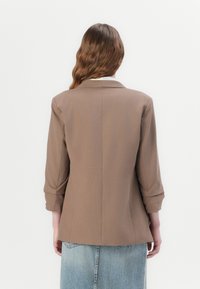 Brun kavaj med strukturerade axlar, trekvartsärmar och en mjuk textur. Bärs över en ljusblå denimkjol.