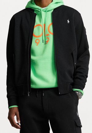 Mann trägt neongrünen Hoodie mit orangefarbenem Text unter schwarzer Reißverschlussjacke und schwarzen Cargohosen, trägt silberne Uhr am linken Handgelenk.