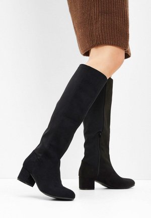 Gambe che indossano stivali alti in suede nero con tacco a blocco, un piede leggermente sollevato, su uno sfondo chiaro e uniforme.