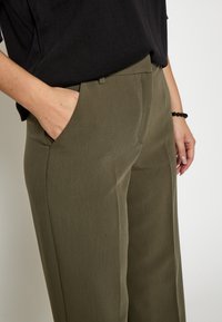 Olijfgroene, op maat gemaakte broek met een recht toegesneden ontwerp, voorzien van zijzakken en een gladde, textuurlijke stof. Gemoed met een zwarte top.