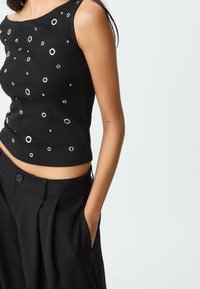 Schwarzes ärmelloses Oberteil mit silbernen Ösen-Details, figurbetonter Schnitt, kombiniert mit hoch taillierten schwarzen Hosen, die eine glatte Textur und einen maßgeschneiderten Sitz zeigen.