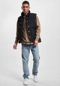 Vest puffer negru cu guler înalt, cu nasturi tip snap, purtat peste un pulover bej cu guler rotund, asortat cu blugi albastri deschis și adidași albi.