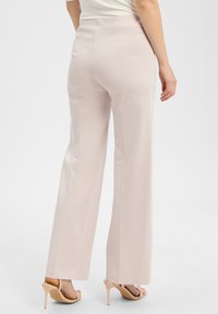 Frau trägt beigefarbene weite Hosen und hautfarbene High Heels, steht vor einem schlichten weißen Hintergrund.