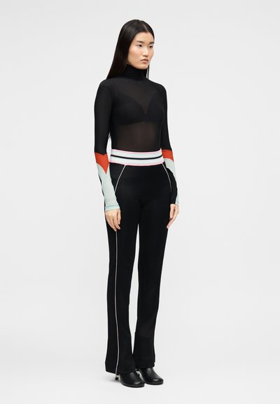 Γυναίκα που στέκεται, φοράει ένα διάφανο μαύρο ζιβάγκο με μανίκια σε έντονα χρώματα, μαύρο παντελόνι με λευκή λεπτομέρεια και μαύρα παπούτσια με τακούνι.