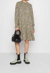 Abito beige con stampa astratta nera, design a strati, maniche lunghe. Borsa a tracolla in pelle nera con parte superiore arricciata. Stivaletti grigio e neri alla caviglia.