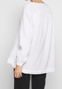 Chemise en lin blanc avec de longues manches légèrement bouffantes, une coupe ample et une surface texturée. L'ourlet est courbé en bas.