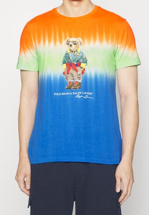 Batikfärgad t-shirt i nyanser av orange, grönt och blått. Fram på tröjan finns en tecknad björn klädd i stilfulla kläder. Kortärmad med rundad halsringning.