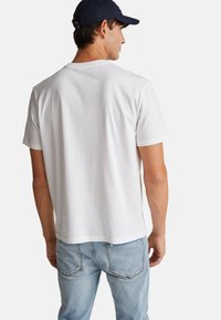 Muž v bílém tričku s krátkým rukávem, světlemodrých džínách a tmavě modré kšiltovce, otočený zády proti jednobarevnému pozadí.