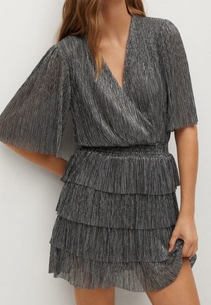 Donna che indossa un abito plissettato grigio argento con maniche corte svolazzanti, scollo a incrocio, vita stretta e gonna a balze a più strati.