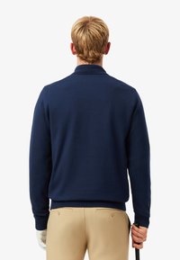 Pull en tricot bleu marine avec col, doté d'une texture lisse et de poignets côtelés. Associé à un pantalon couleur crème et un club de golf noir.