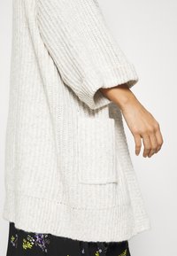 Cardigan in maglia chunky di un beige chiaro. Presenta maniche arrotolate e una tasca a toppa sul lato. Texture morbida con righe orizzontali.