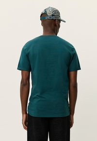 Τυρκουάζ βαμβακερό T-shirt με στρογγυλή λαιμόκοψη και κοντά μανίκια, φορεμένο με μαύρο κοντό παντελόνι με σχέδια, φωτογραφημένο από πίσω.