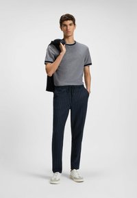 Gray short-sleeve t-shirt with navy trim, paired with navy pinstripe trousers and white sneakers, held jacket in one hand.