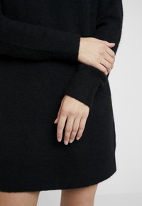 Abito in maglia nera con una texture morbida, polsini a coste e orlo arrotondato. Ingrandimento di mani che poggiano sul tessuto.