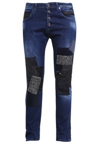 Vaqueros de mezclilla en azul oscuro con forma ajustada. Presenta múltiples parches texturizados en negro y gris, incluyendo materiales con lentejuelas y tejidos.